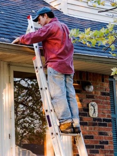 A man on an aluminum ladder clearing the gutter along the roofline of a brick house.