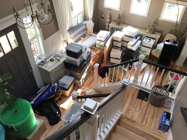 A living room seen from a staircase landing, filled with stacked moving boxes, plastic bins and packing paper.
