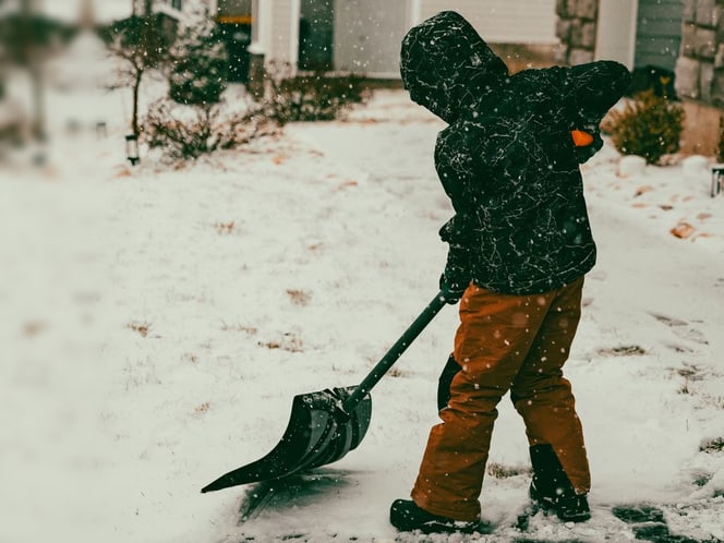 Someone in a hooded winter coat shoveling a snowy walkway while snow falls