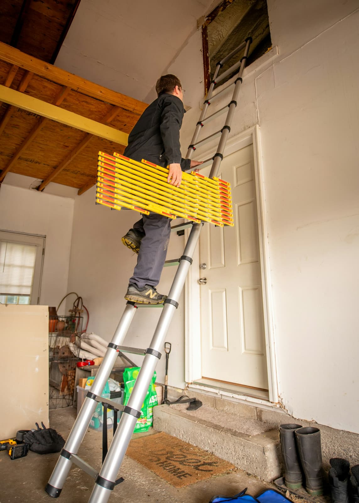A man seen from behind on a ladder in a garage, working at the attic hatch above the door.