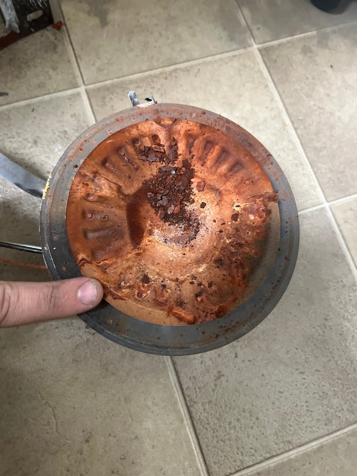A corroded water heater part crusted with rust and sediment, held over a tile floor.