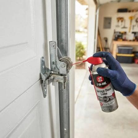 A gloved hand spraying lubricant onto the roller and hinge of a garage door.
