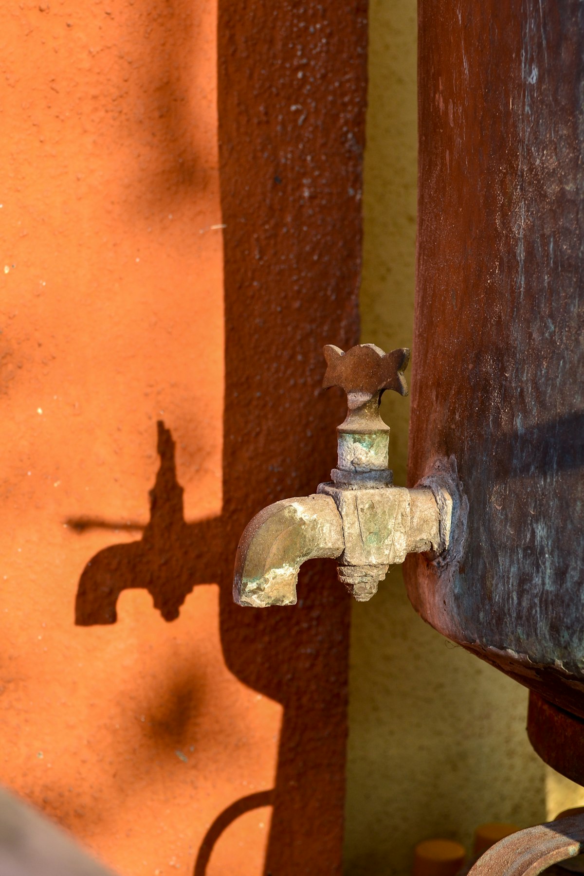 An old brass tap on the side of a rusted metal tank, its shadow cast on a sunlit orange wall.