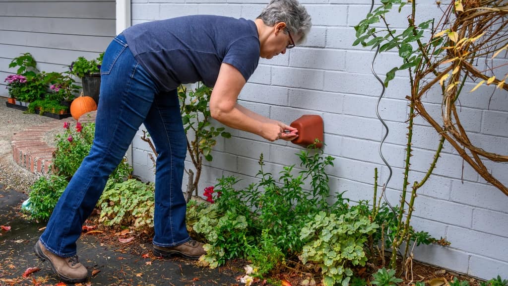 A woman bending to check the dryer vent hood on the outside wall of a house, garden beds at her feet.