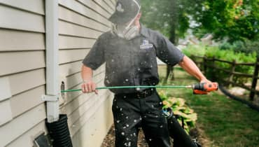 A technician in a mask running a rotary brush into a dryer vent on the side of a house.