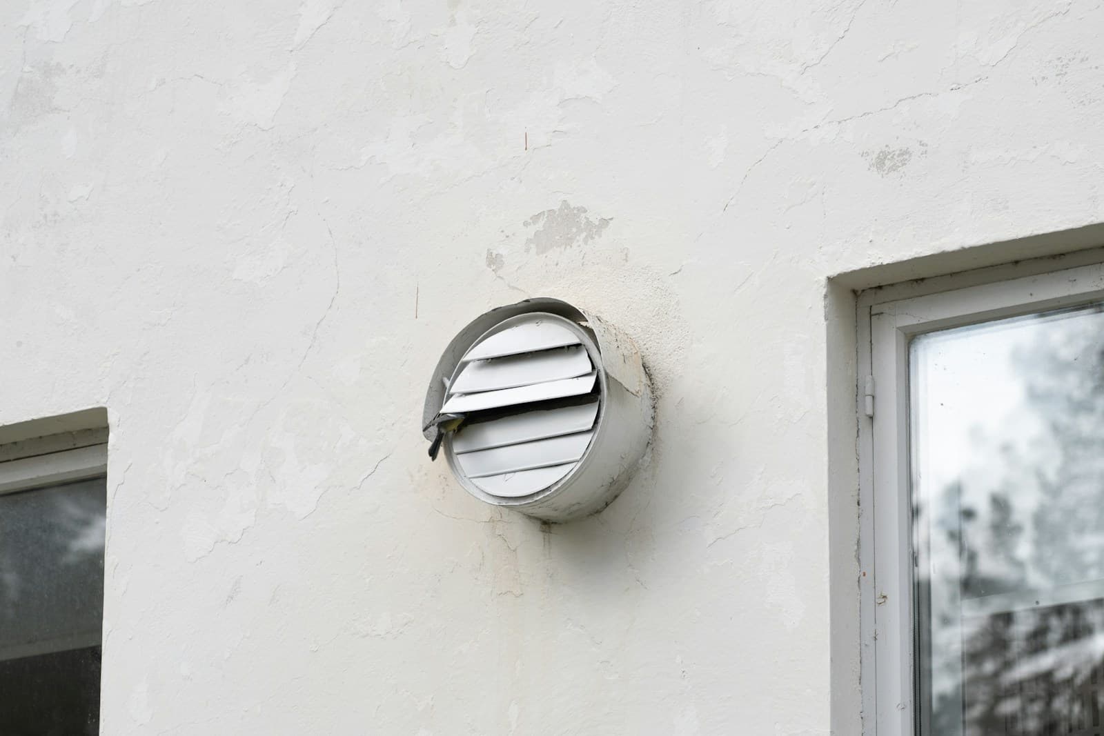 A clear louvered dryer vent hood on a white exterior wall between two windows.
