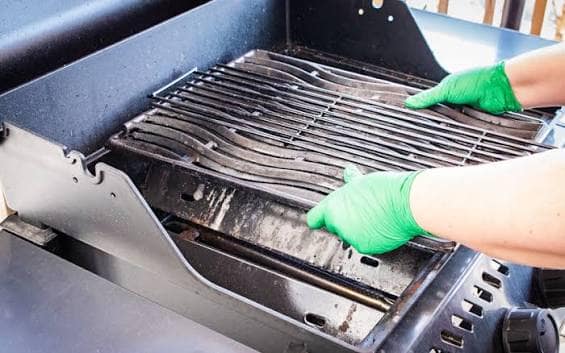 Gloved hands lifting the cooking grates out of a gas grill for cleaning.