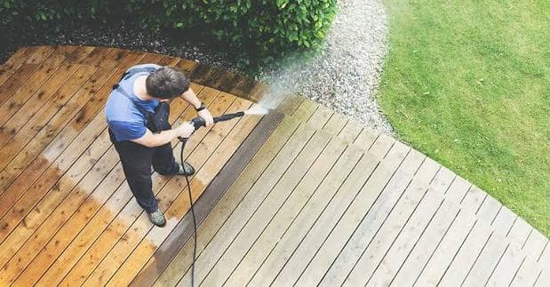 A man pressure washing a wooden deck, a clean stripe showing through the gray grime.