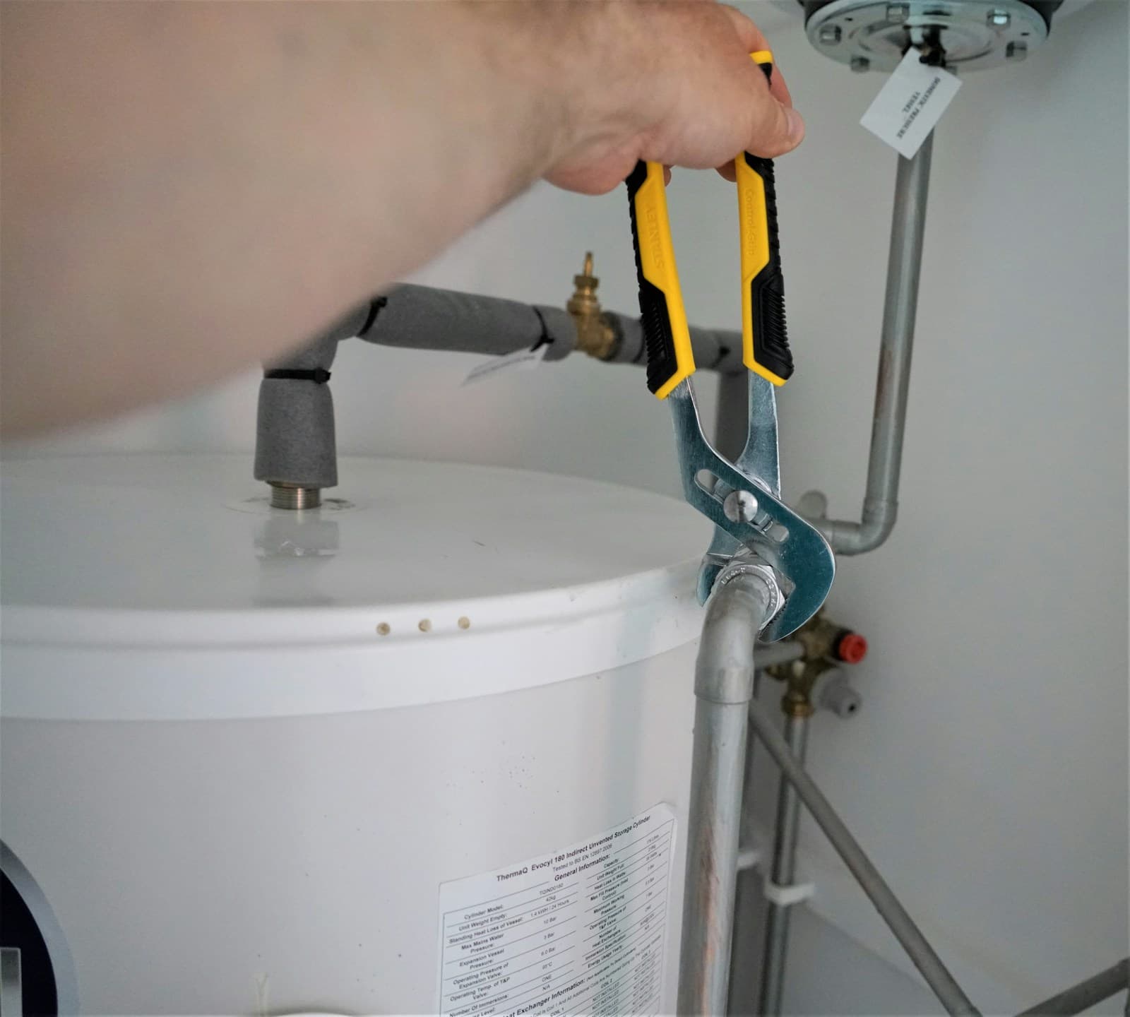 Hands tightening the water line fitting on top of a white tank water heater with a pair of pliers.