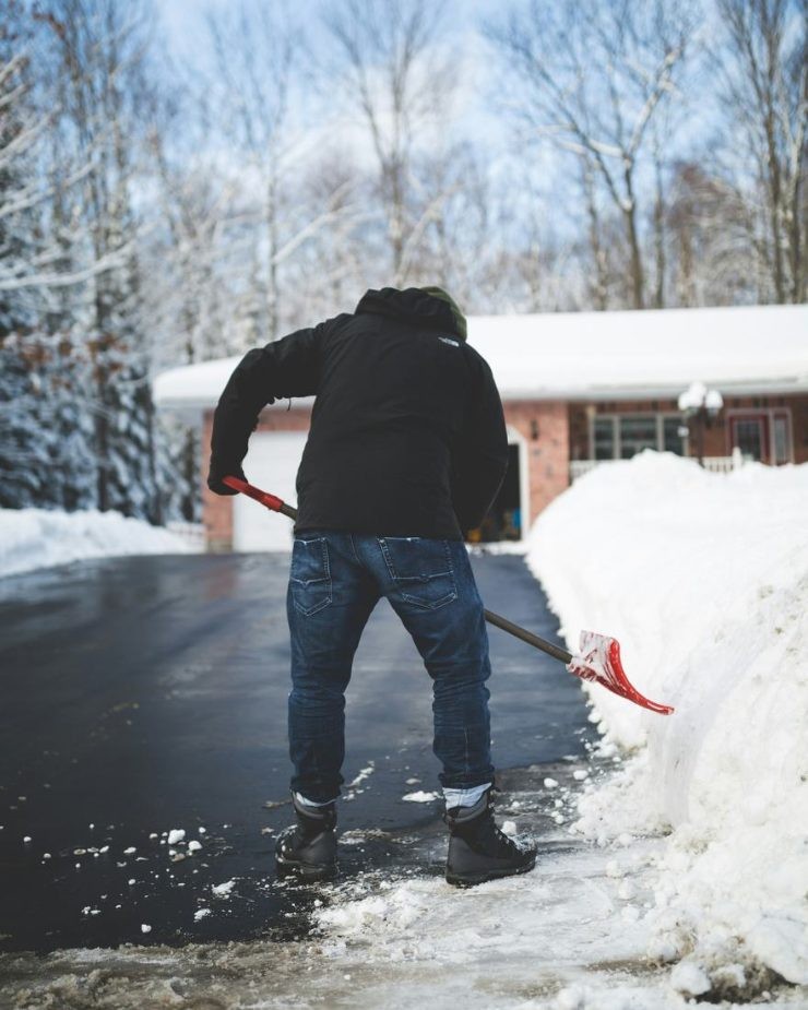 A man in a black jacket shoveling snow off a driveway in front of a brick ranch house.